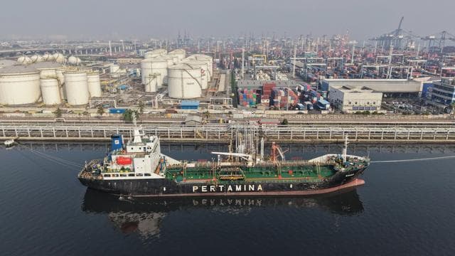 A large ship branded "Pertamina" alongside a dock with a tank farm and shipping containers